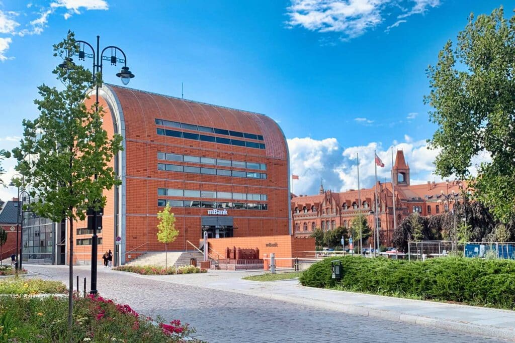 A modern, brick-red warehouse-style building at the mBank headquarters in Bydgoszcz, with the historic Poczta Gł&oacute;wna in the background